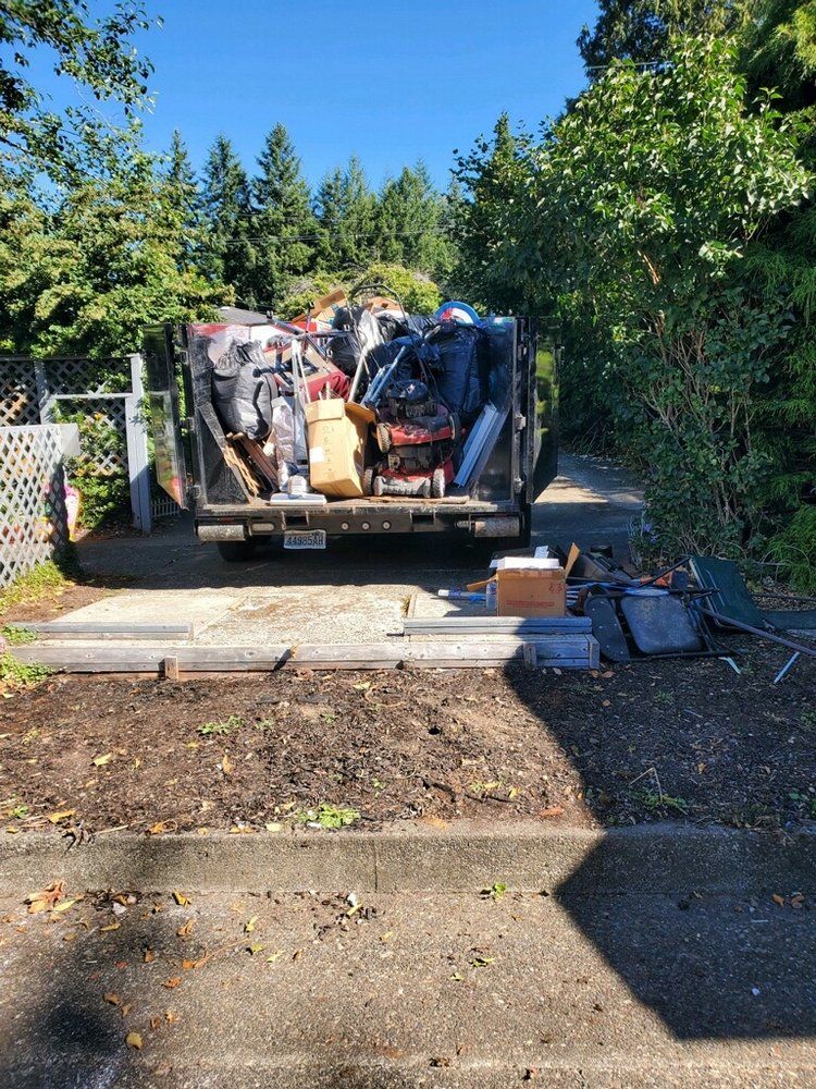 A truck overflowing with junk parked on a driveway; debris scattered nearby; trees in the background.