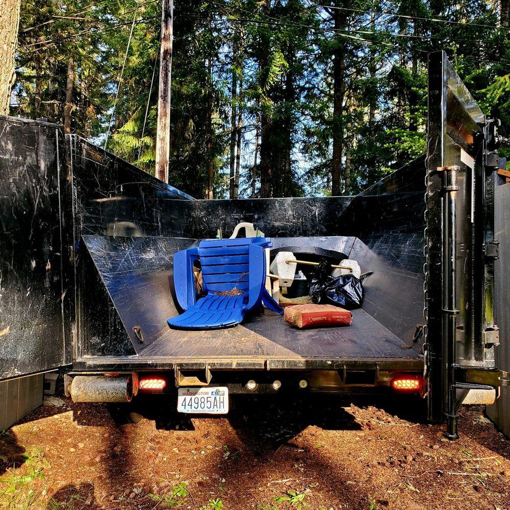 A blue lawn chair, engine, and items sit in a black dump truck bed in a forest.