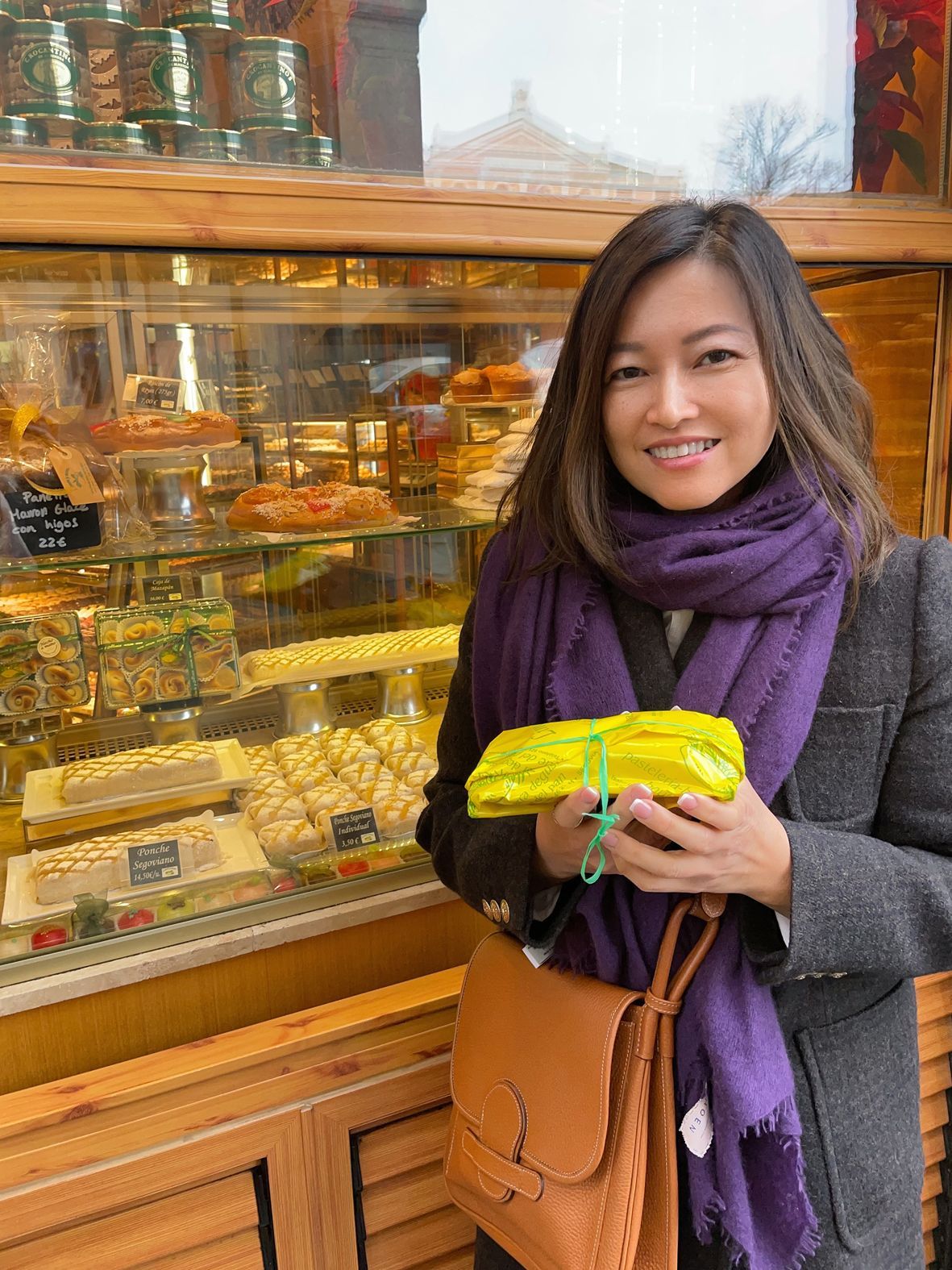A woman in a purple scarf is holding a bag of yellow flowers in front of a bakery.