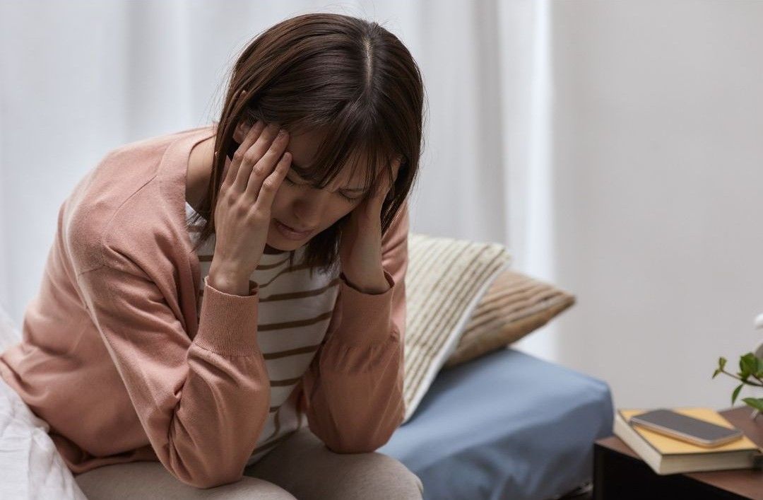 A person sitting indoors with their head in their hands, appearing distressed or experiencing a headache.