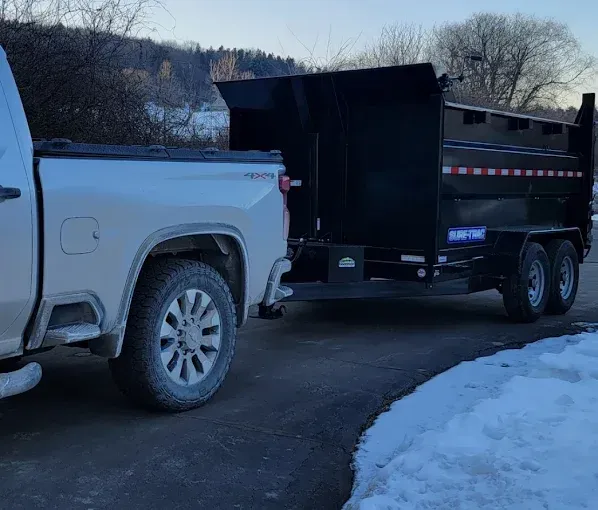 Silver pickup truck towing a black dump trailer on a snowy driveway.