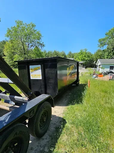 Black dumpster on a trailer parked on a dirt path next to a grassy area on a sunny day.