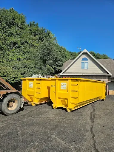 Two large yellow dumpsters sit on a driveway, a truck trailer is nearby. Behind is a house and trees, under a blue sky.