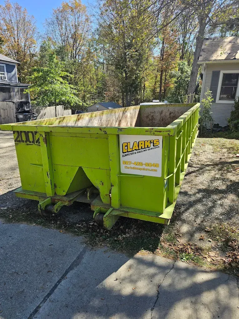 A lime-green roll-off dumpster from Clark's Dumpster Service sits on a paved driveway in front of a house and trees.