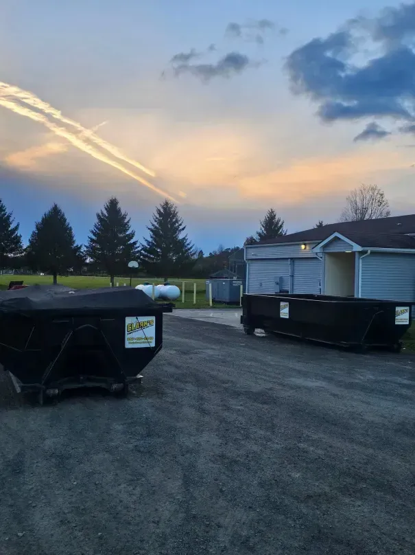 Two black dumpsters on gravel with trees and a building at sunset.