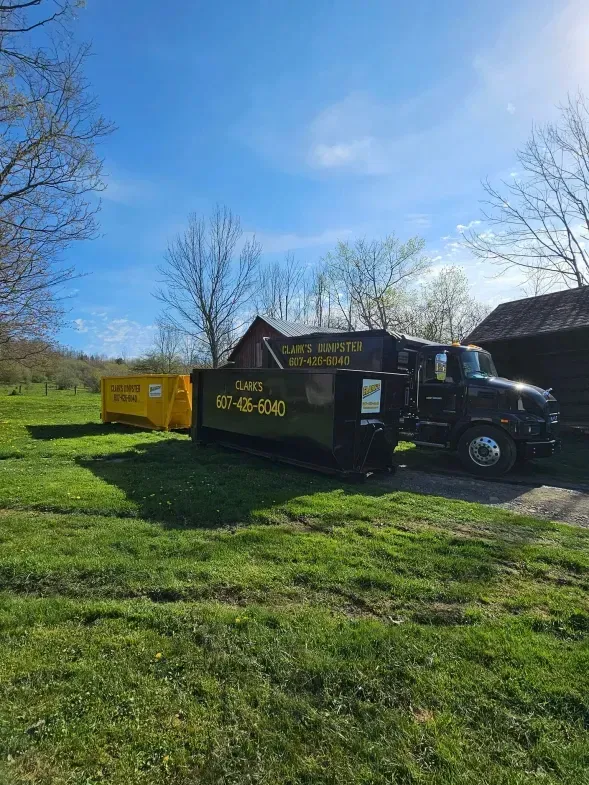 Dump truck with two dumpsters on grassy land under a blue sky. One dumpster is yellow, the other is black.