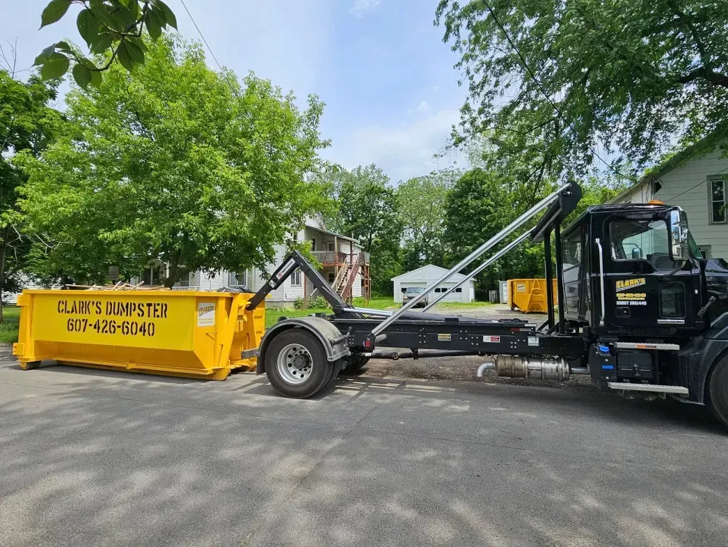 Yellow dumpster being lifted by a black roll-off truck on a driveway, with trees and a house in the background.