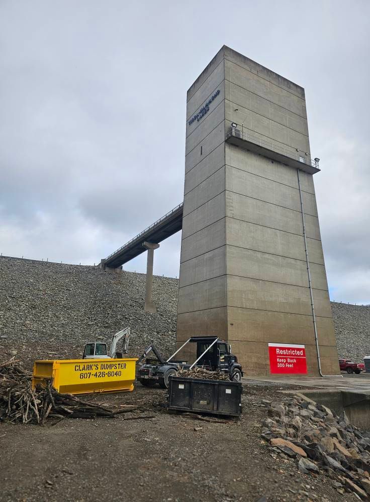 A large concrete intake tower stands next to a rock-filled dam with construction equipment and dumpsters at the base.