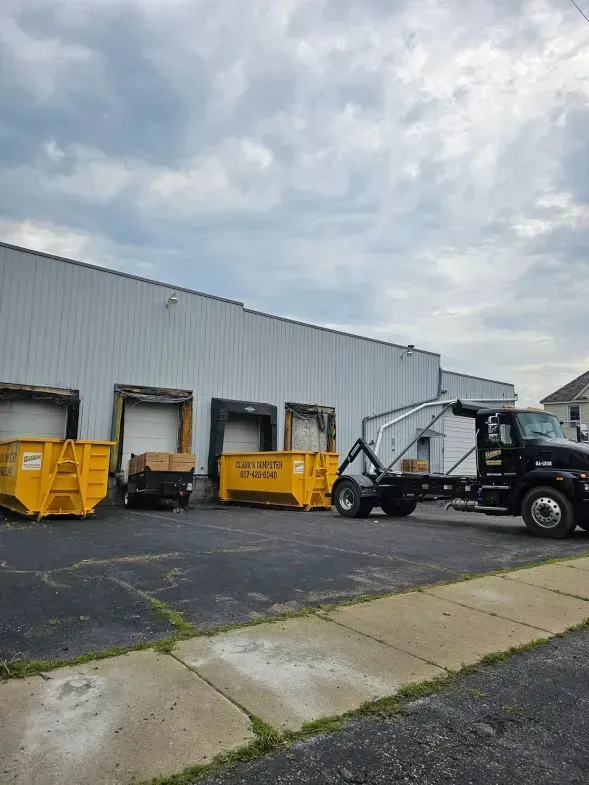 Dumpster truck near a warehouse loading yellow dumpsters, under a cloudy sky.