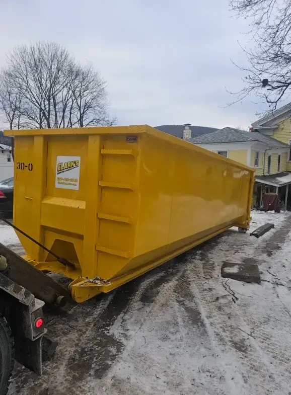 Yellow dumpster on a snowy road, behind a truck. Houses and bare trees are in the background.