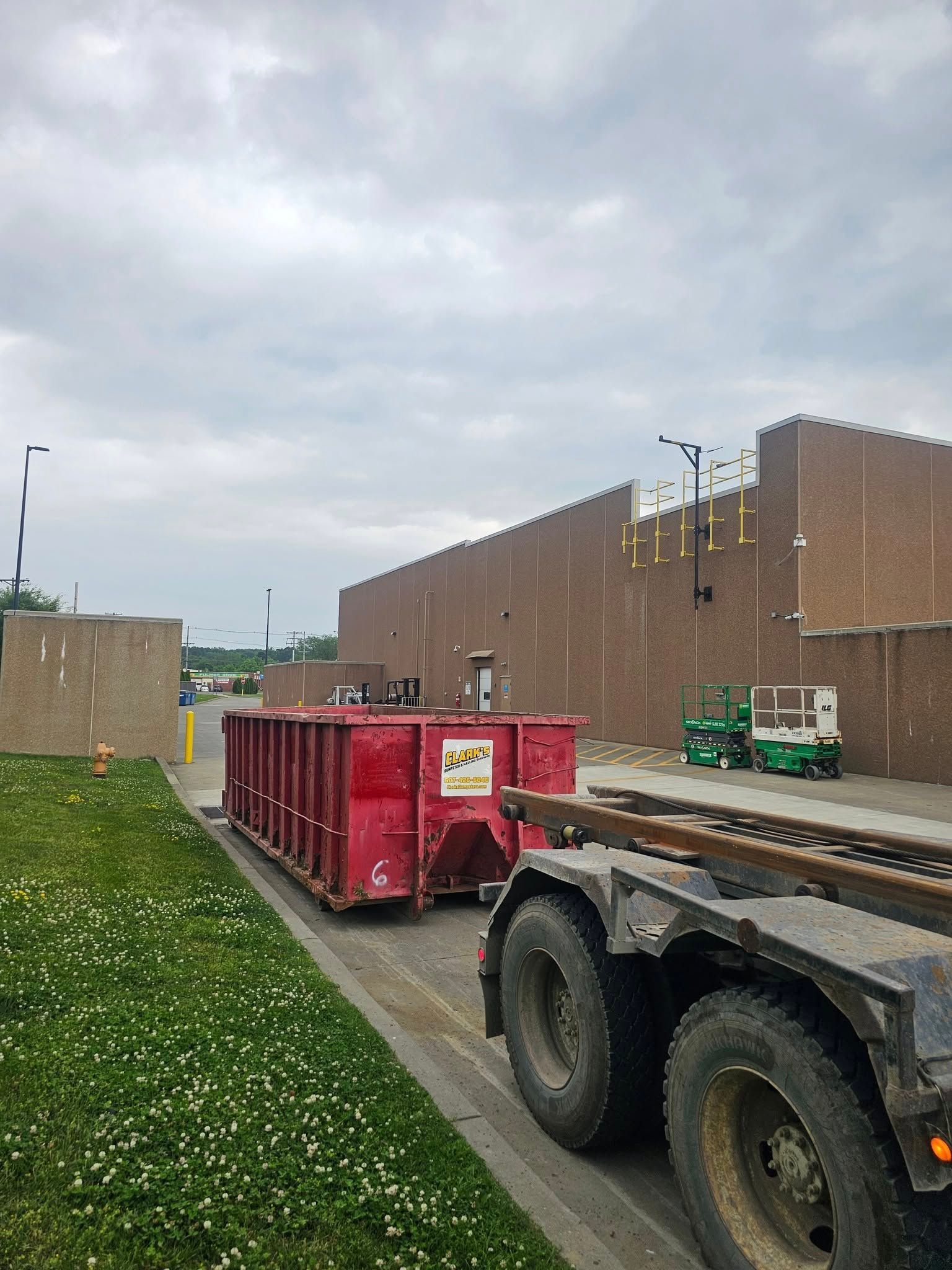 A red industrial dumpster sits next to a truck in a grassy construction area beside a brick building under a cloudy sky.