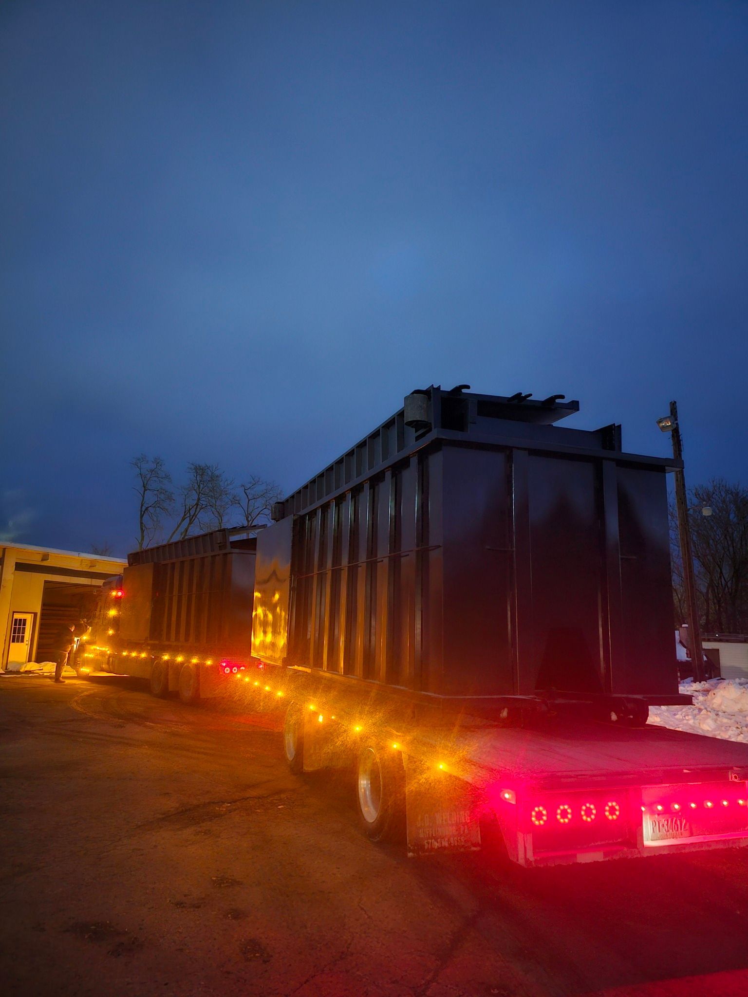 A large, dark metal dumpster sits on a trailer at dusk, illuminated by glowing orange side lights and red tail lights.