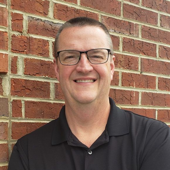 Man with glasses smiling in front of a brick wall, wearing a black collared shirt.