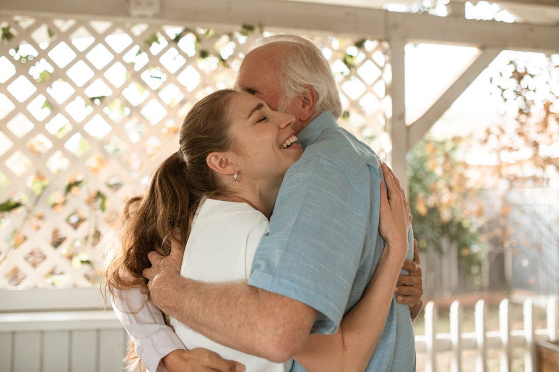 Two people hugging warmly in a sunlit garden gazebo with a white lattice backdrop.