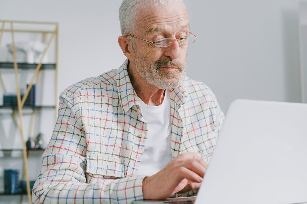 Older person with glasses, using a laptop, in a well-lit room, typing with a focused expression.