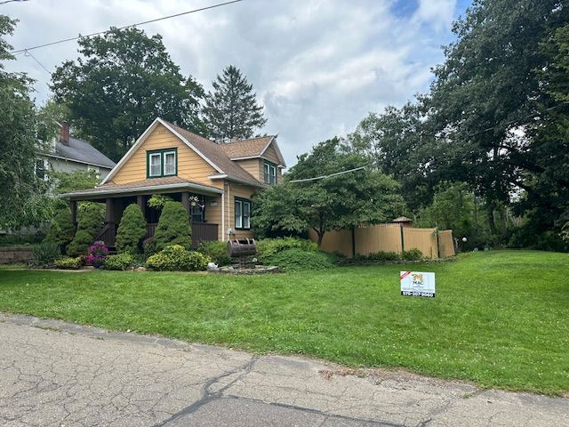 Yellow house with porch and lawn, under a cloudy sky. Sign on the lawn.