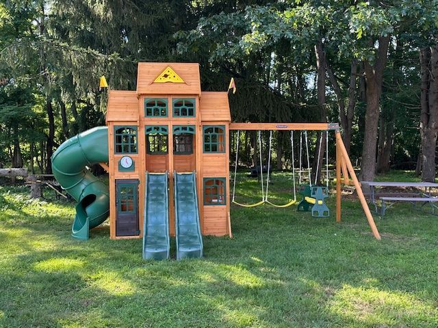 Wooden playset in a yard with a slide, swings, and a fort-like structure. Green slide, brown wood, green grass.
