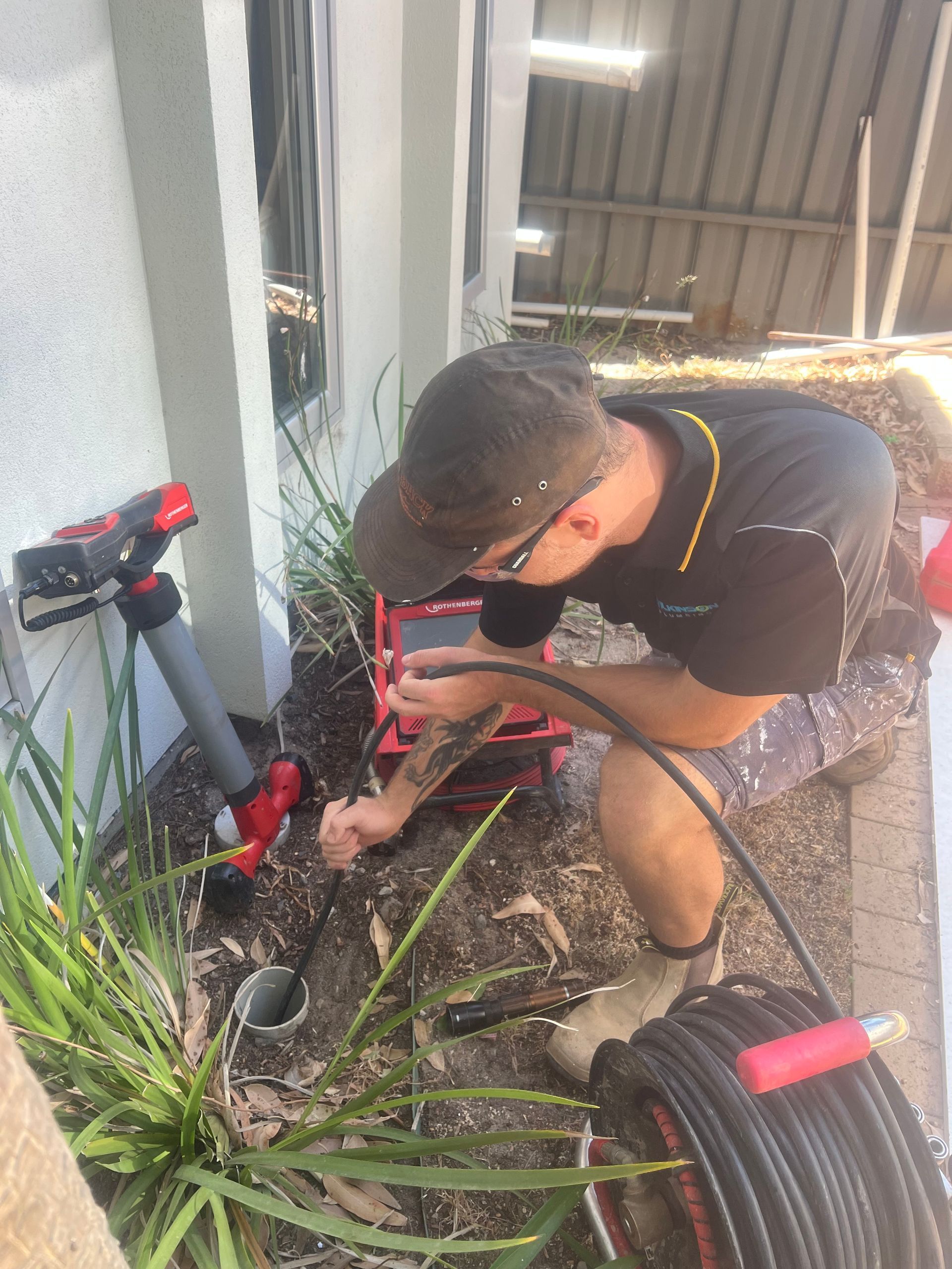 Plumber inspecting a drain with a camera. Red equipment, gray pipes, and green plants are nearby.