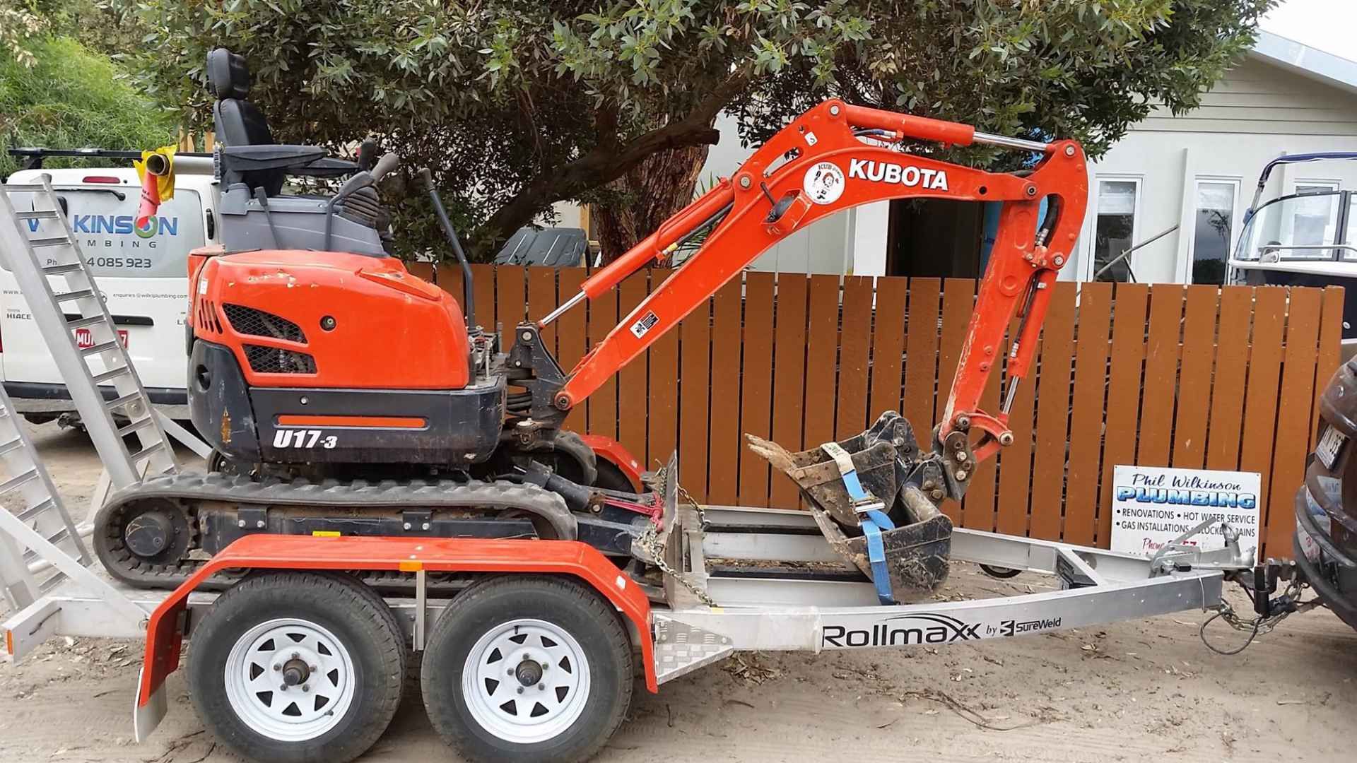 Orange Kubota mini excavator on a trailer parked next to a brown wooden fence.