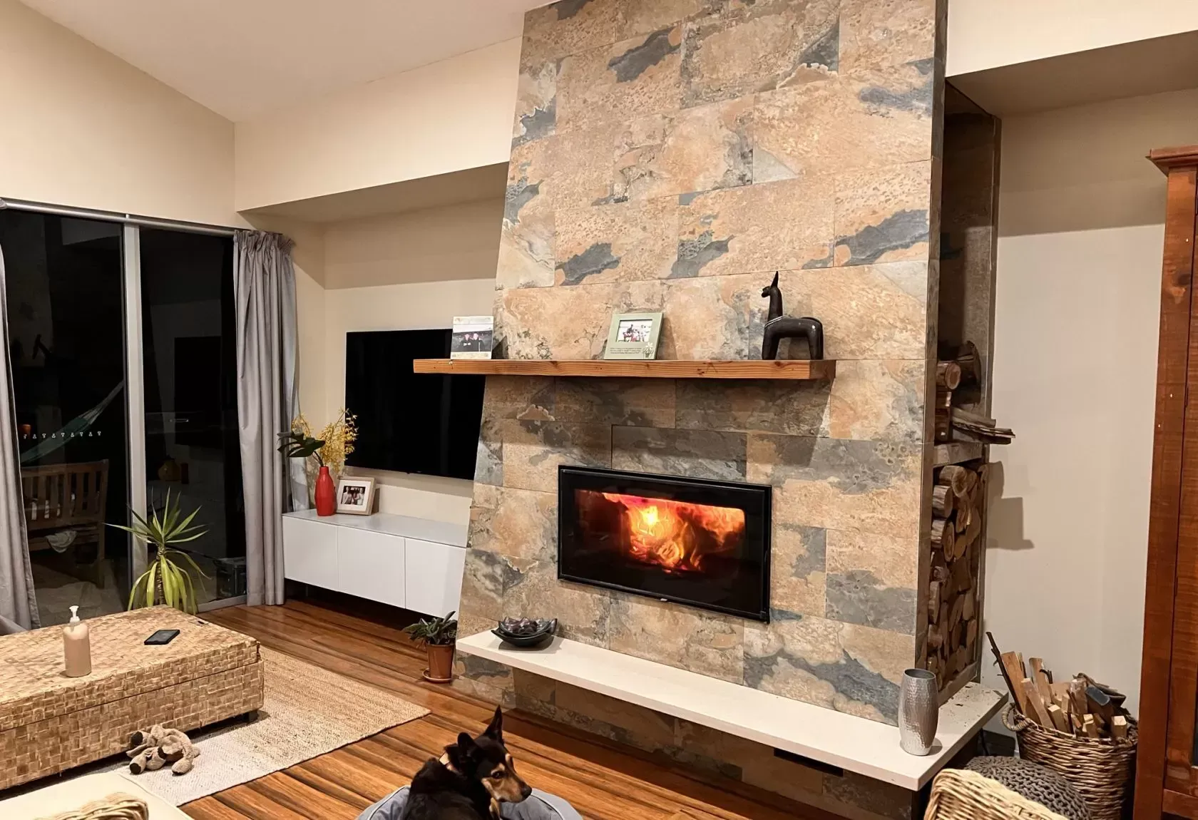 Living room with fireplace, TV, and dog. Fire burning in stone fireplace; a dog resting on the floor.