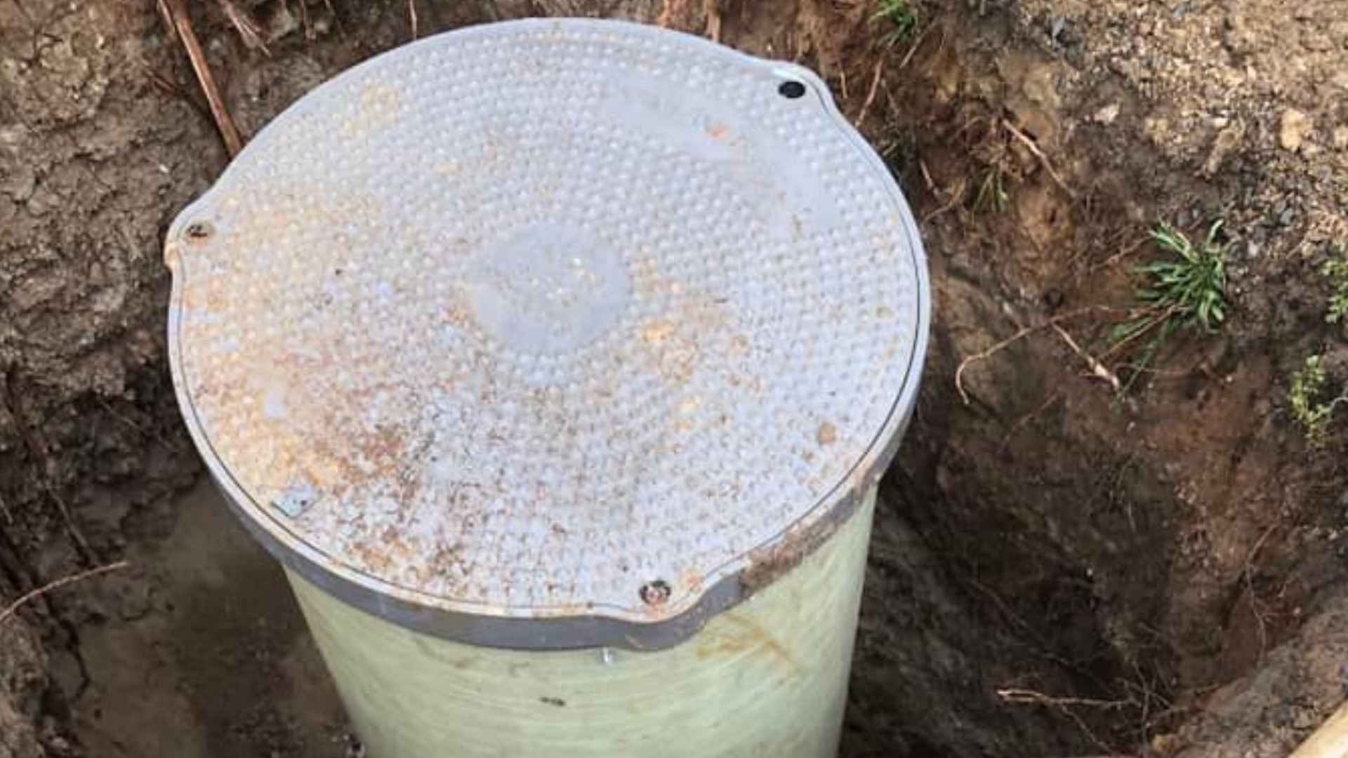 A round, gray lid covering a buried, cylindrical utility structure in a dirt trench.