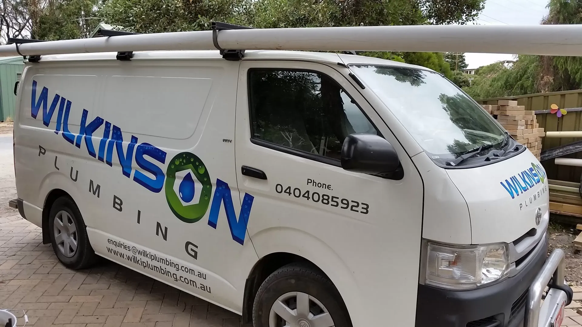 White van with "Wilkinson Plumbing" logo, parked on a street. Roof racks hold white pipes.