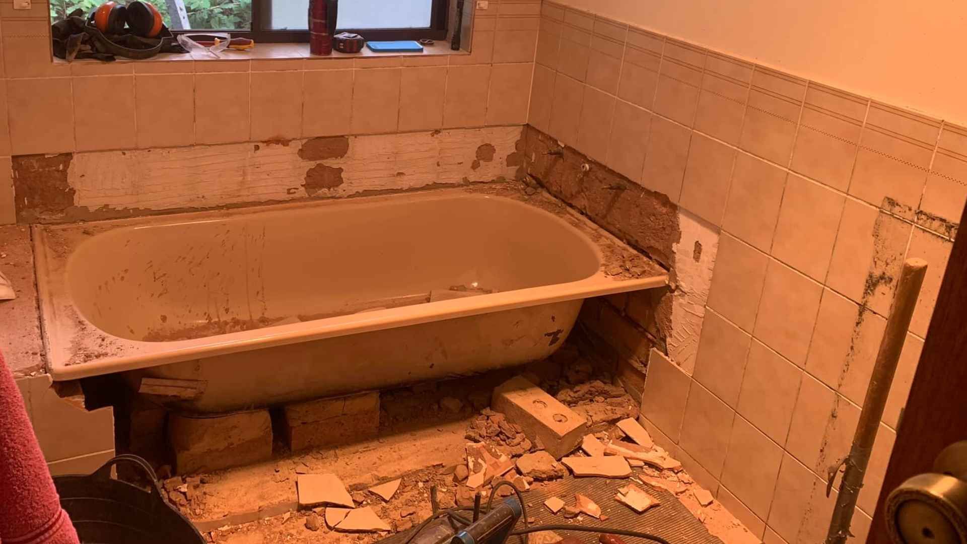 Bathroom renovation in progress; tub and wall tiles partially removed, debris on the floor, bricks supporting the tub.