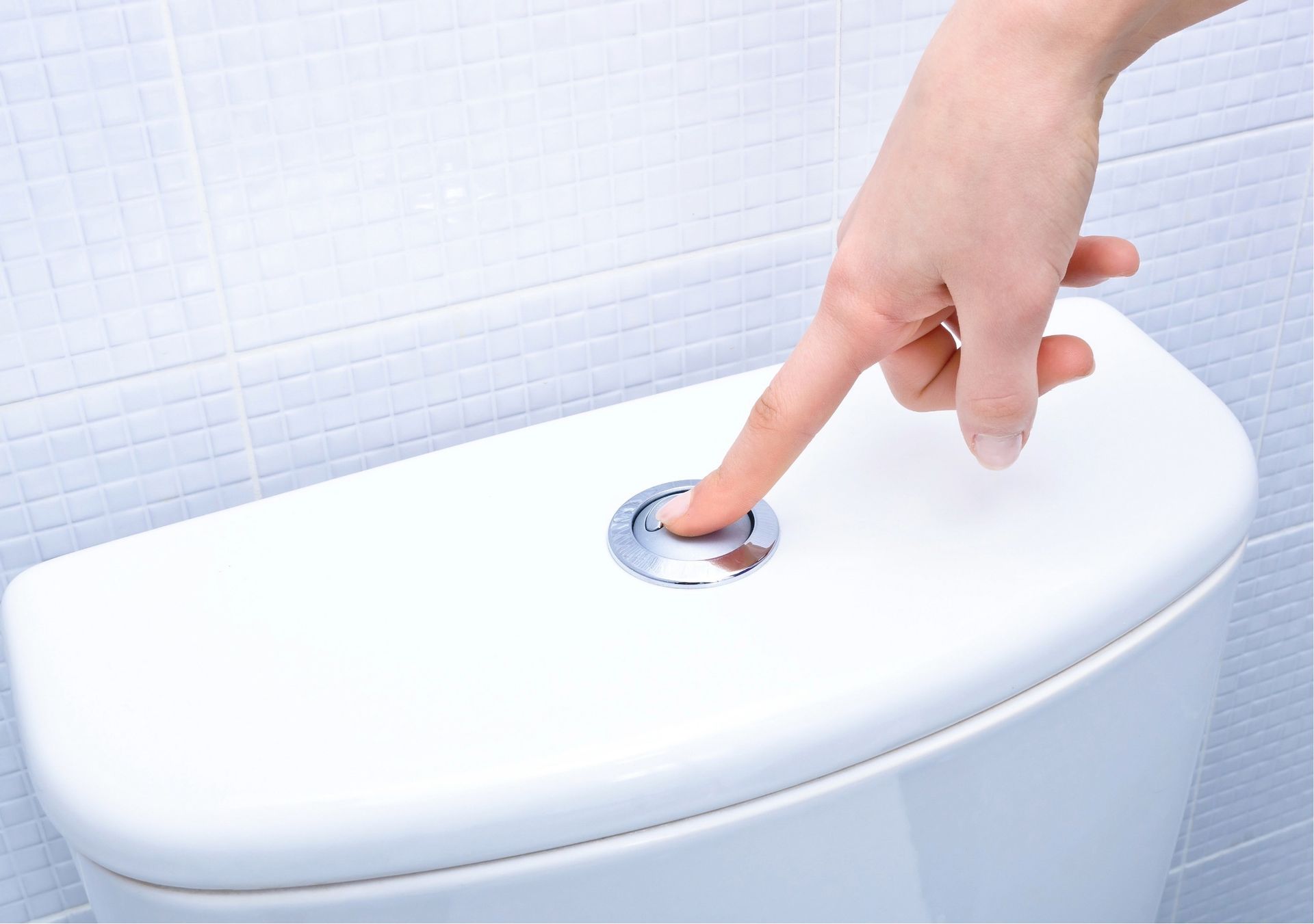 Hand pressing a silver flush button on a white toilet tank in a bathroom with tiled walls.