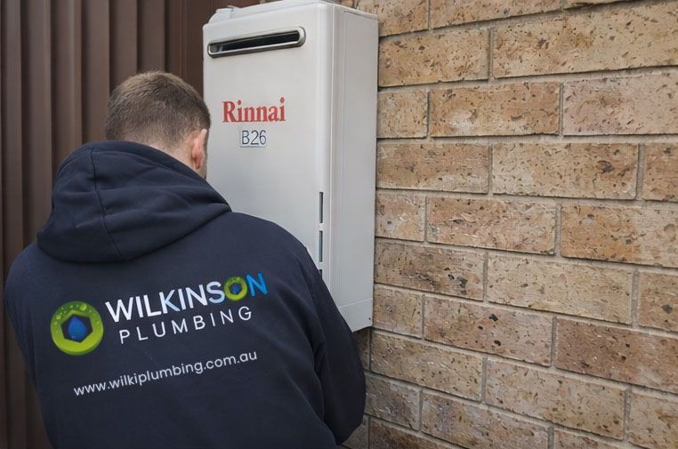 Plumber in blue sweatshirt installing a Rinnai water heater on a brick wall.