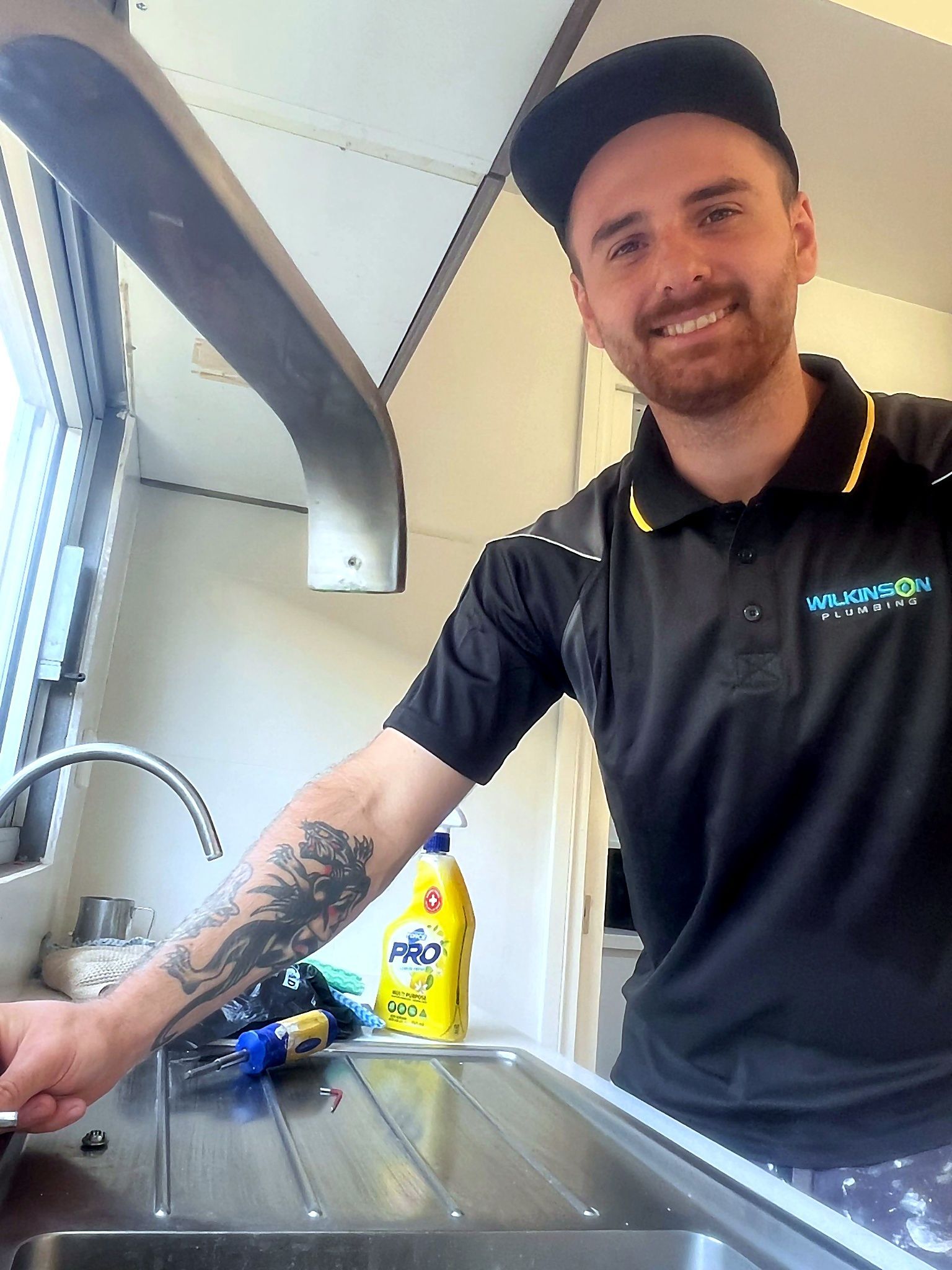 Man in a black polo shirt, black cap, smiling, beside a kitchen sink with a tattoo on his arm.