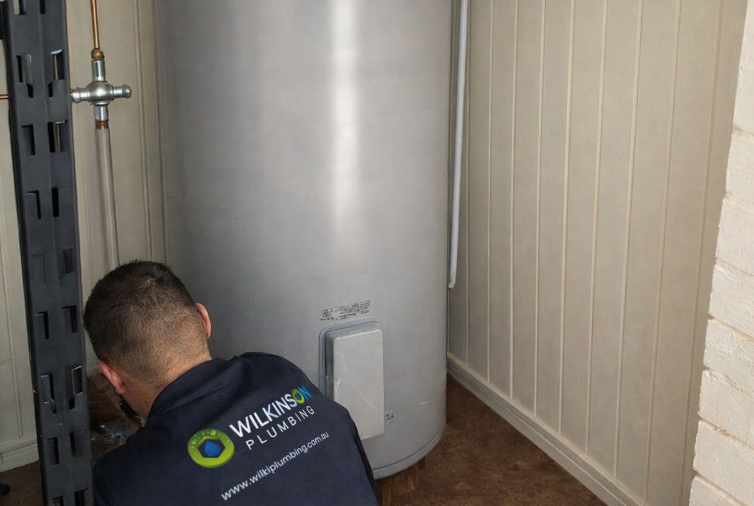 Plumber working on a water heater inside a utility room. Grey tank, white walls, plumber wearing a blue shirt.