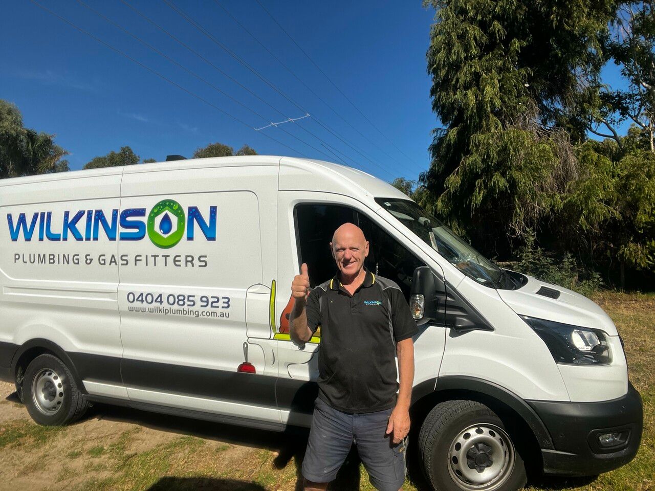 A man gives a thumbs-up in front of a white Wilkinson Plumbing & Gas Fitters work van parked outdoors.