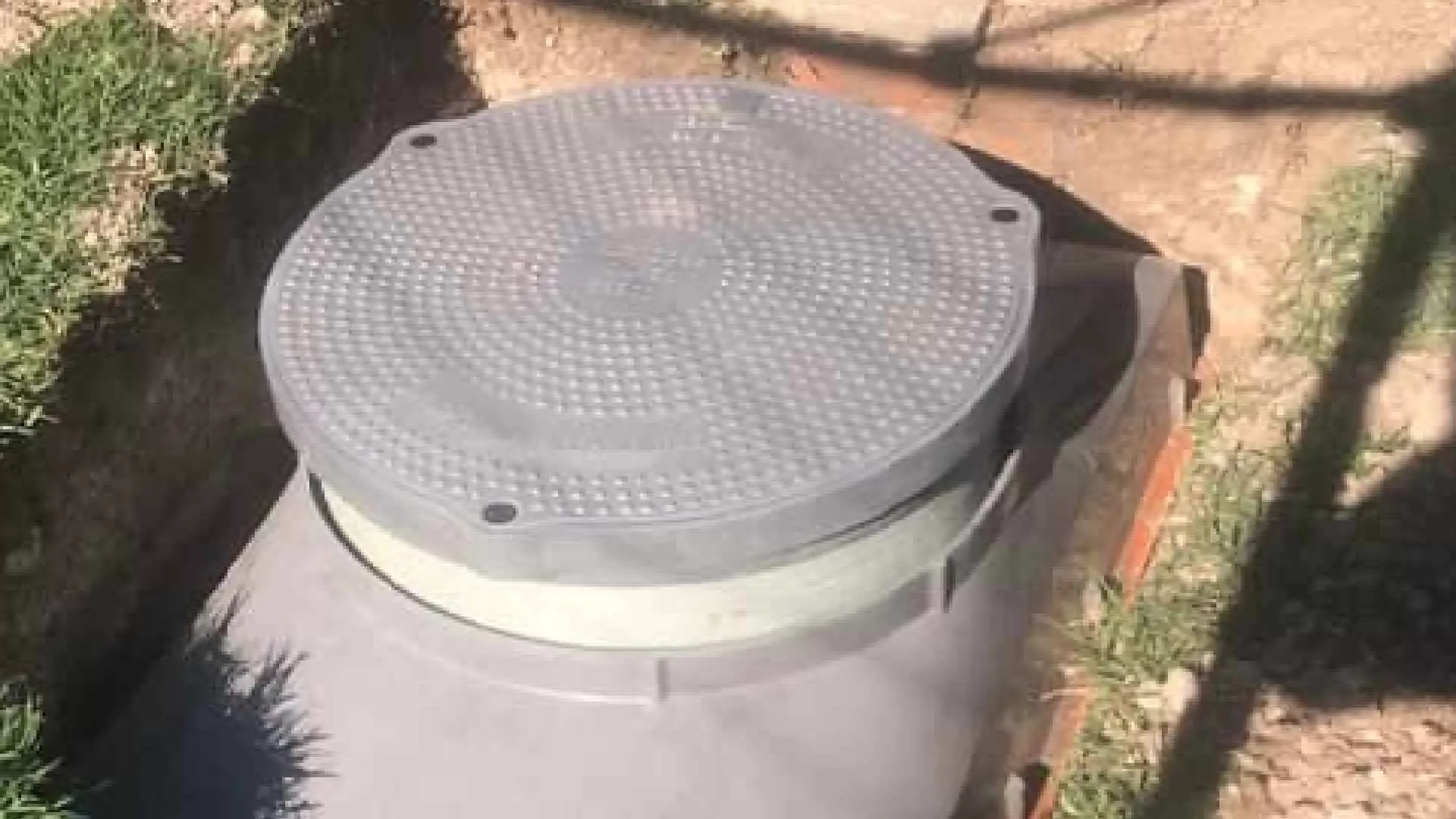 Gray, round manhole cover on a cylindrical structure, partially buried in the ground. Surrounded by grass and a brick border.