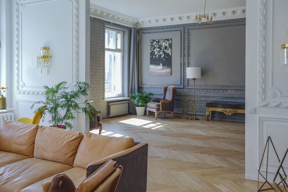 Sala de estar con sofá de cuero color canela, pisos de madera, plantas y paredes grises y blancas.