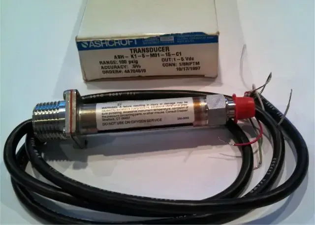 Transparent cylindrical electronic component with attached black cable and a small labeled box on a white surface