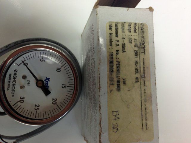 Close-up of a round pressure gauge beside a beige cardboard box on a white surface.