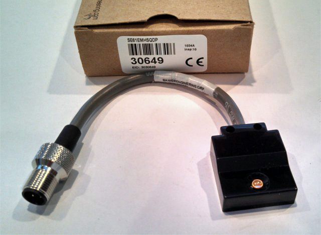 Black cable adapter with metal connector and small box, shown in front of its packaging.