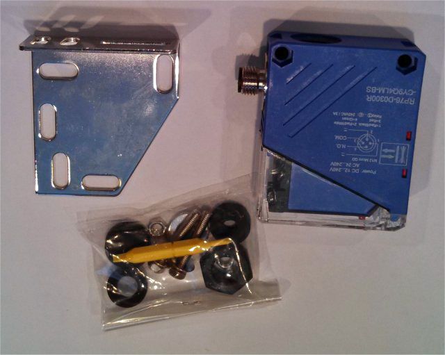 Blue industrial sensor, metal mounting bracket, and small parts in plastic bags on a white surface