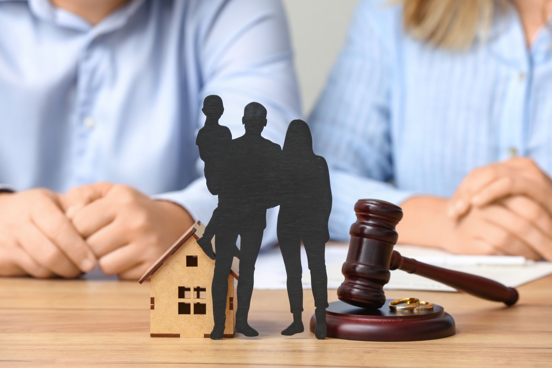 Silhouettes of a family beside a small house model, gavel, and rings on a table.