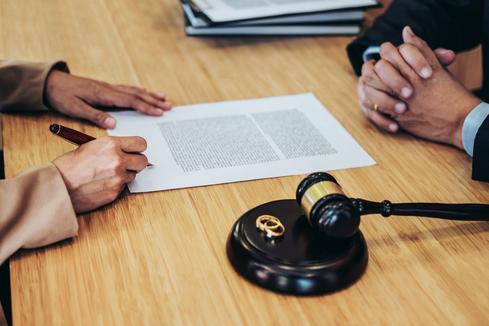 A person signs a legal document at table while another listens, with gavel and wedding rings nearby