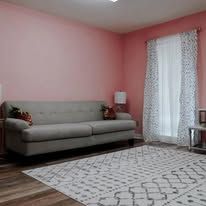 Pink room with gray couch, white curtains, and patterned rug.