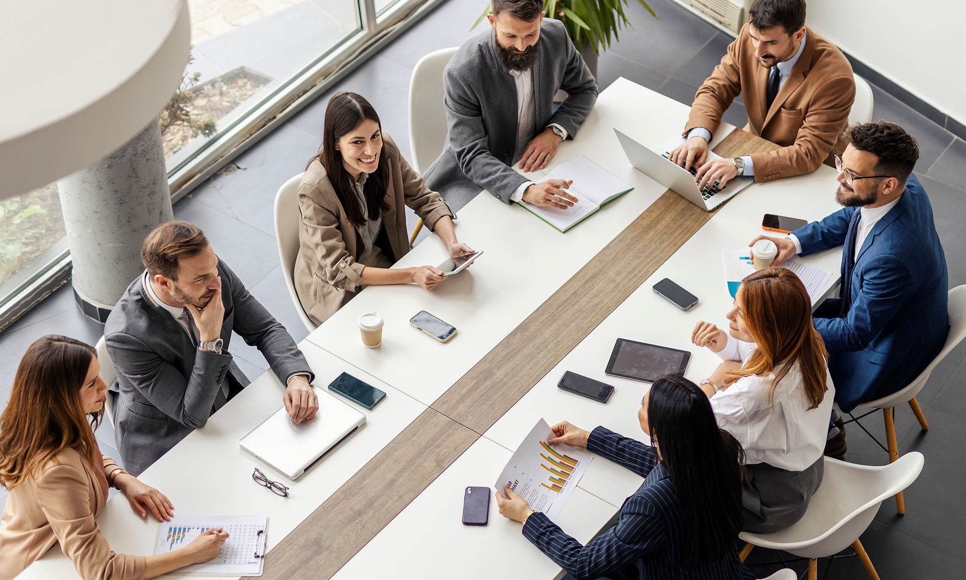 People in business attire at a conference table with laptops and papers, discussing.