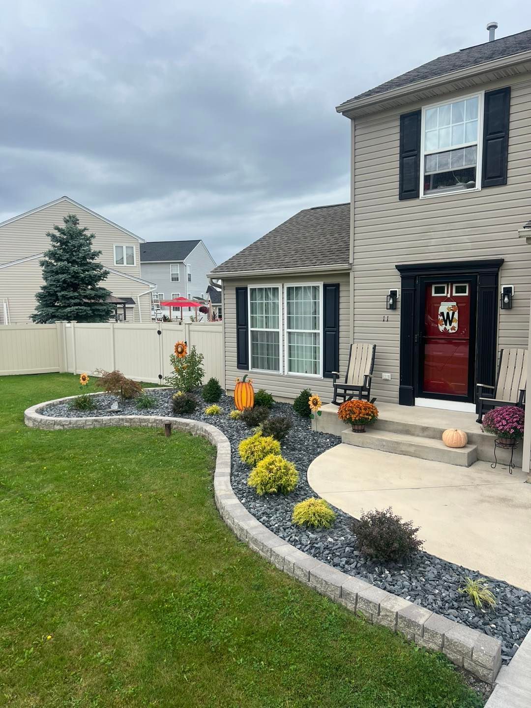 A two-story beige house with a curved flower bed filled with plants and pumpkins.