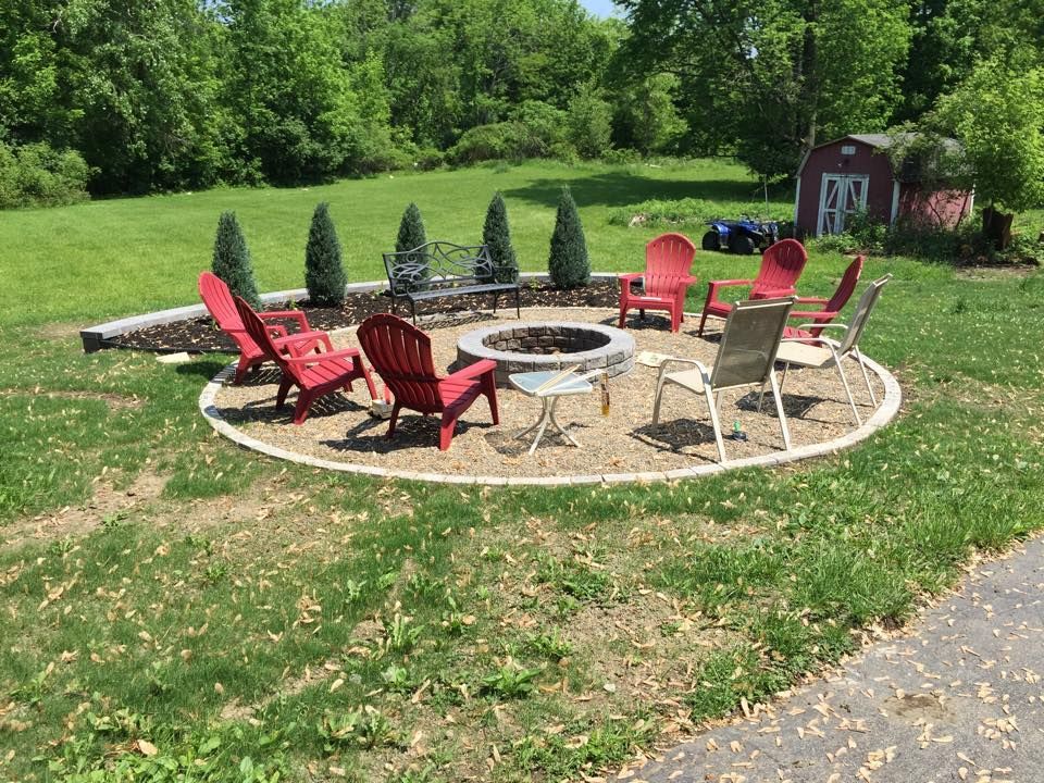 A circular fire pit area with red and neutral-colored chairs, surrounded by greenery and trees.