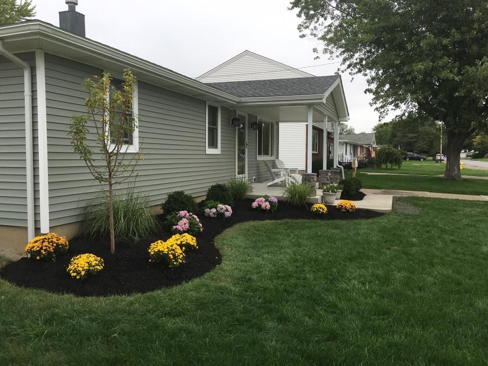 House exterior with manicured lawn and flower bed containing yellow, pink, and orange mums and a small tree.