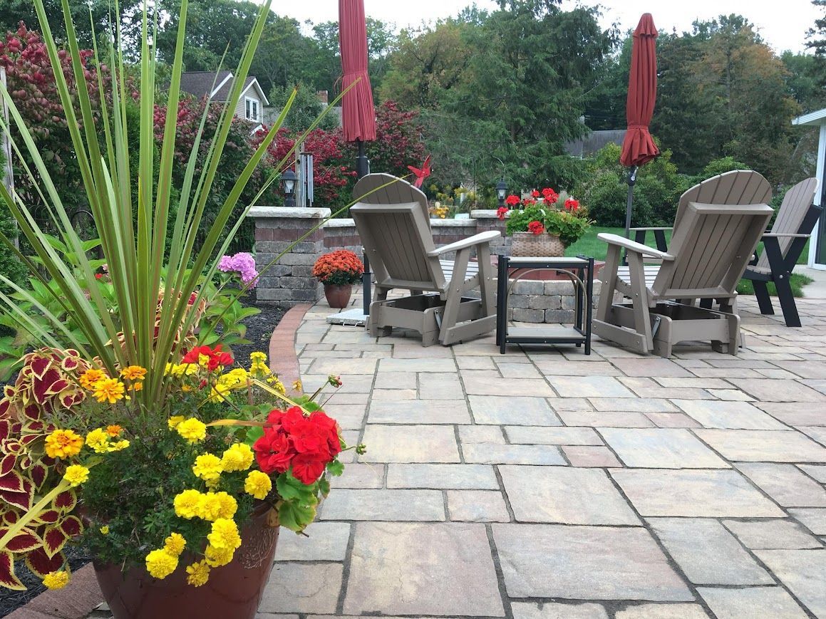 Patio with stone pavers, Adirondack chairs, red umbrellas, and potted flowers.