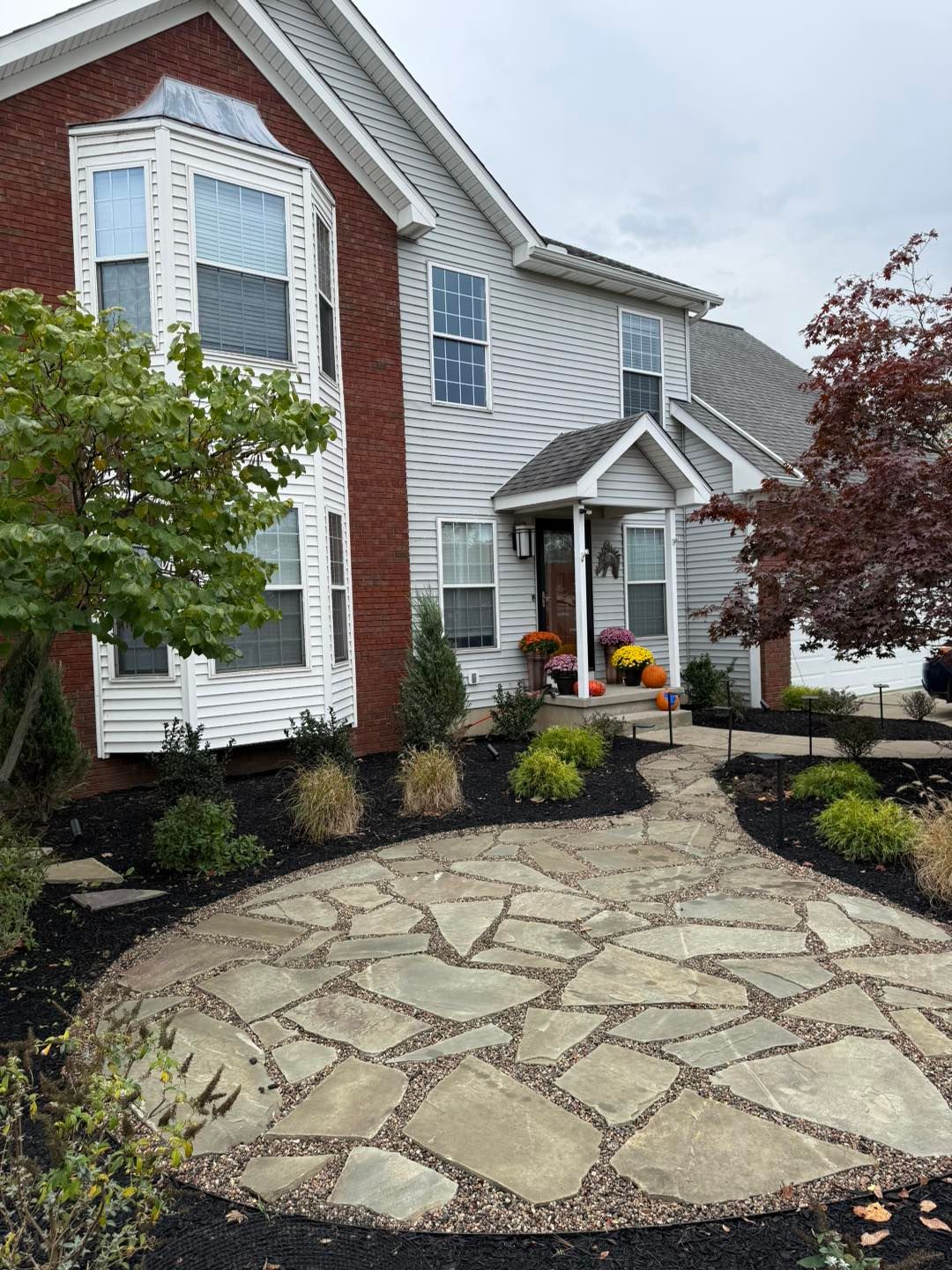 Stone path leading to a two-story house with brick and gray siding. Autumn decorations on porch.
