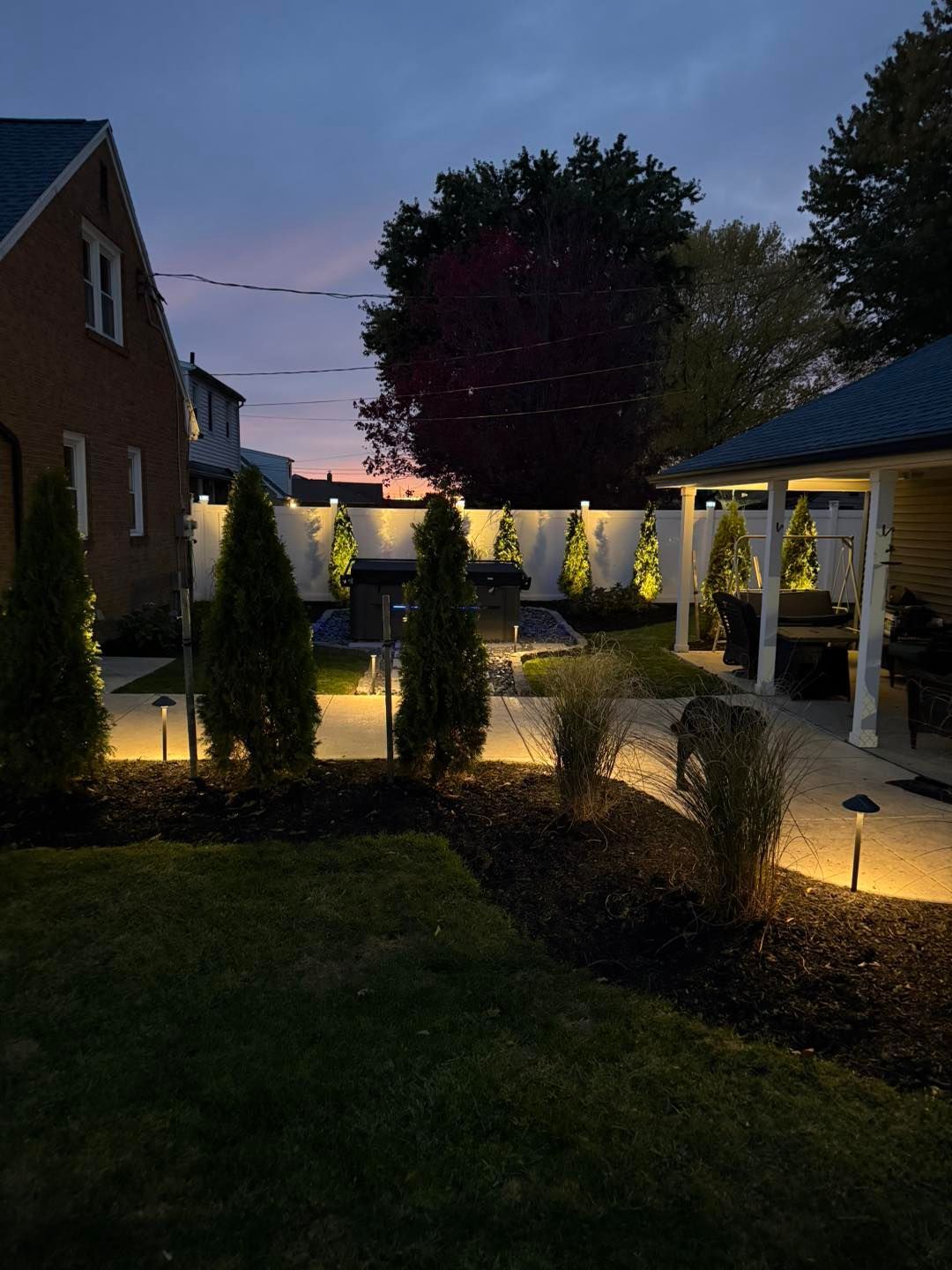 Backyard at dusk with landscape lighting illuminating trees, a white wall, and a patio.