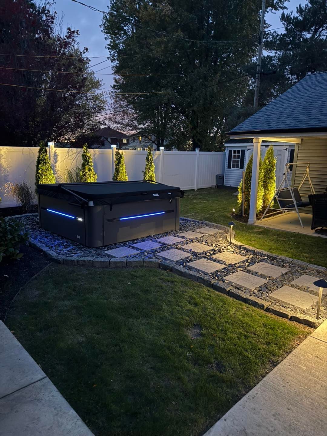 Backyard with lit hot tub, stone pathway, lawn, and surrounding trees, at dusk.