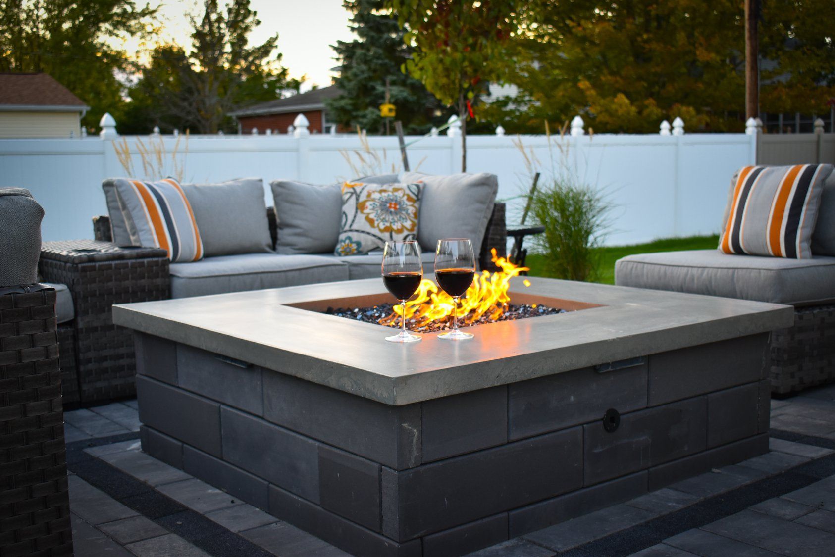 Outdoor seating area with lit fire pit and wine glasses. Gray patio furniture, white fence, evening light.